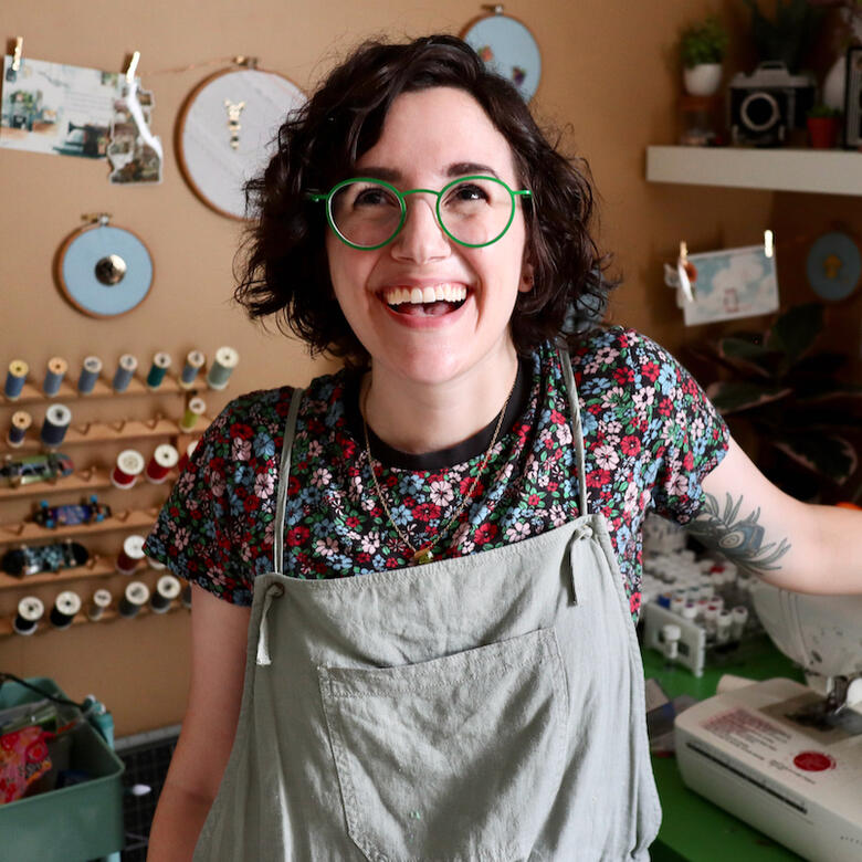Lauren is sitting in their studio with a happy grin on their face and one arm resting on a sewing machine on the table behind them. They're wearing bright green, round glasses.